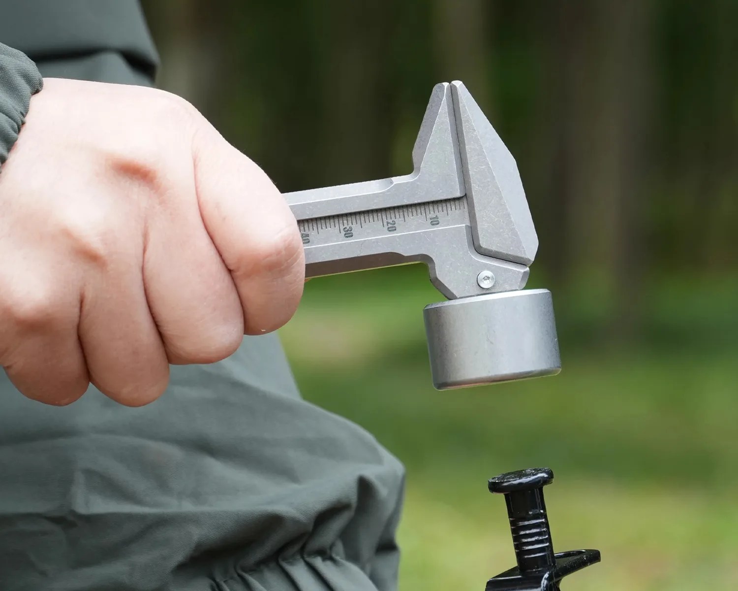 Hand holding a silver caliper measuring a black metal object outdoors.