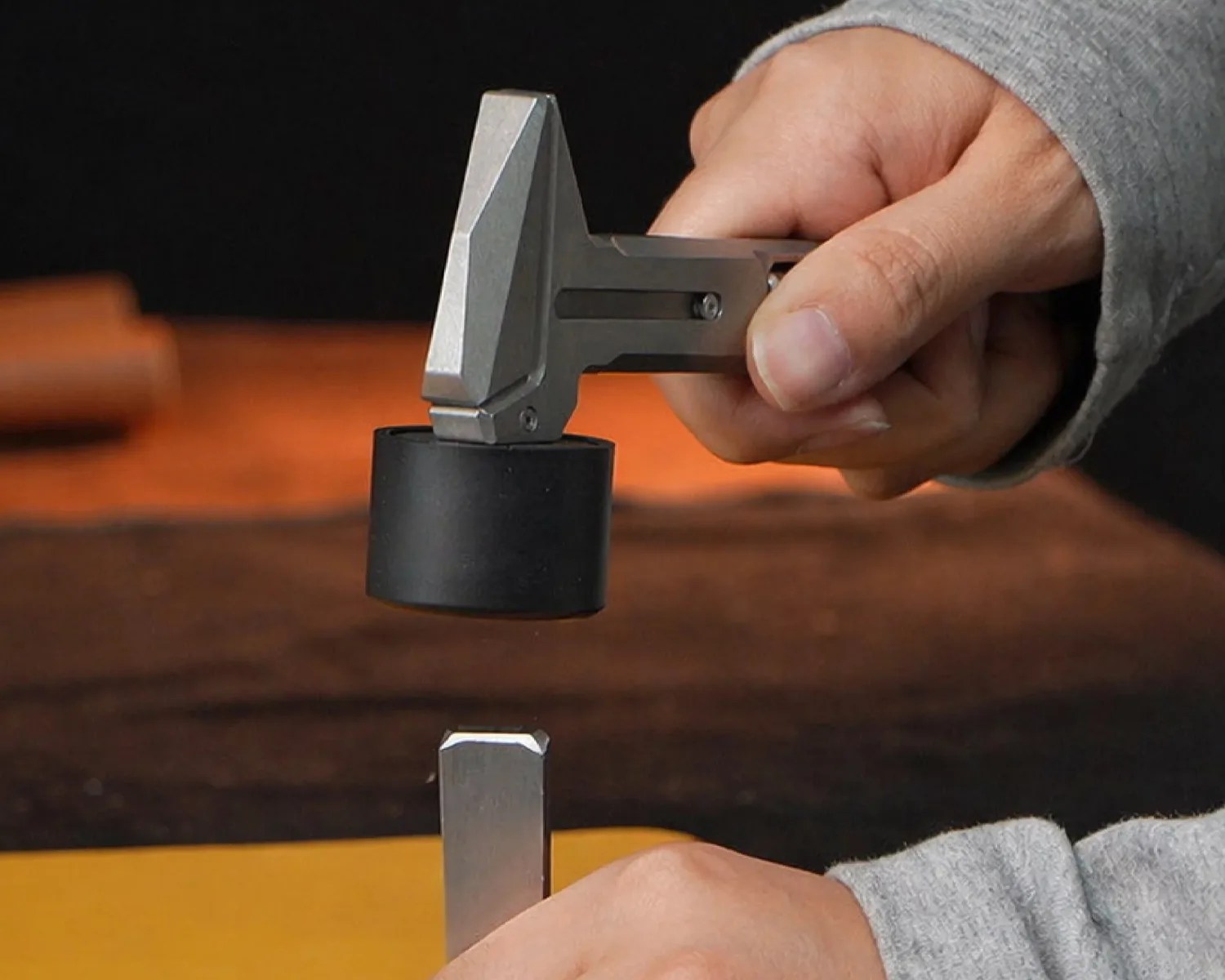 Hand holding a small metal hammer with a black rubber head above a metal chisel on a wooden surface.