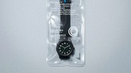 Black analog wristwatch with white numbers and green second hand in clear plastic packaging.