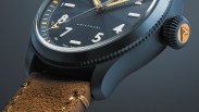 Close-up of a black watch with white numerals, gold hands, textured brown leather strap, and a black crown with a gold emblem.