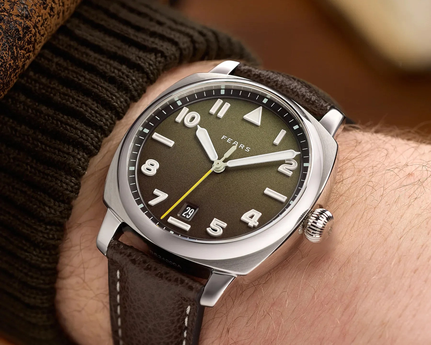 Silver wristwatch with olive green dial, white numerals, yellow second hand, and brown leather strap worn on a wrist.