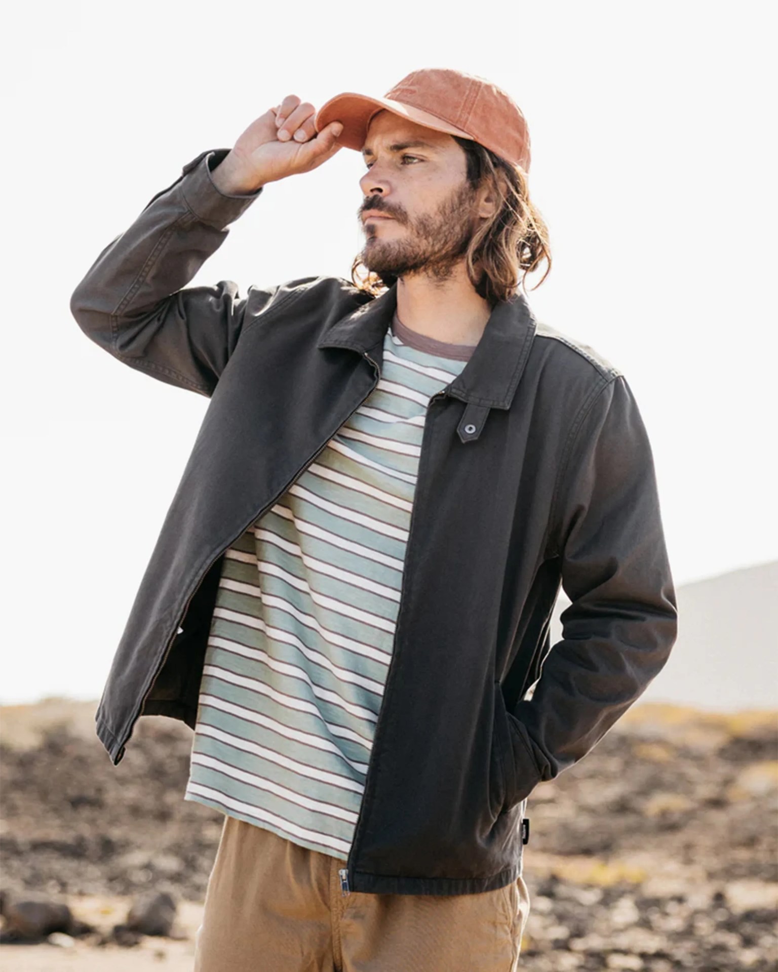 Man wearing a faded black jacket, striped shirt, tan pants, and a faded orange cap outdoors.