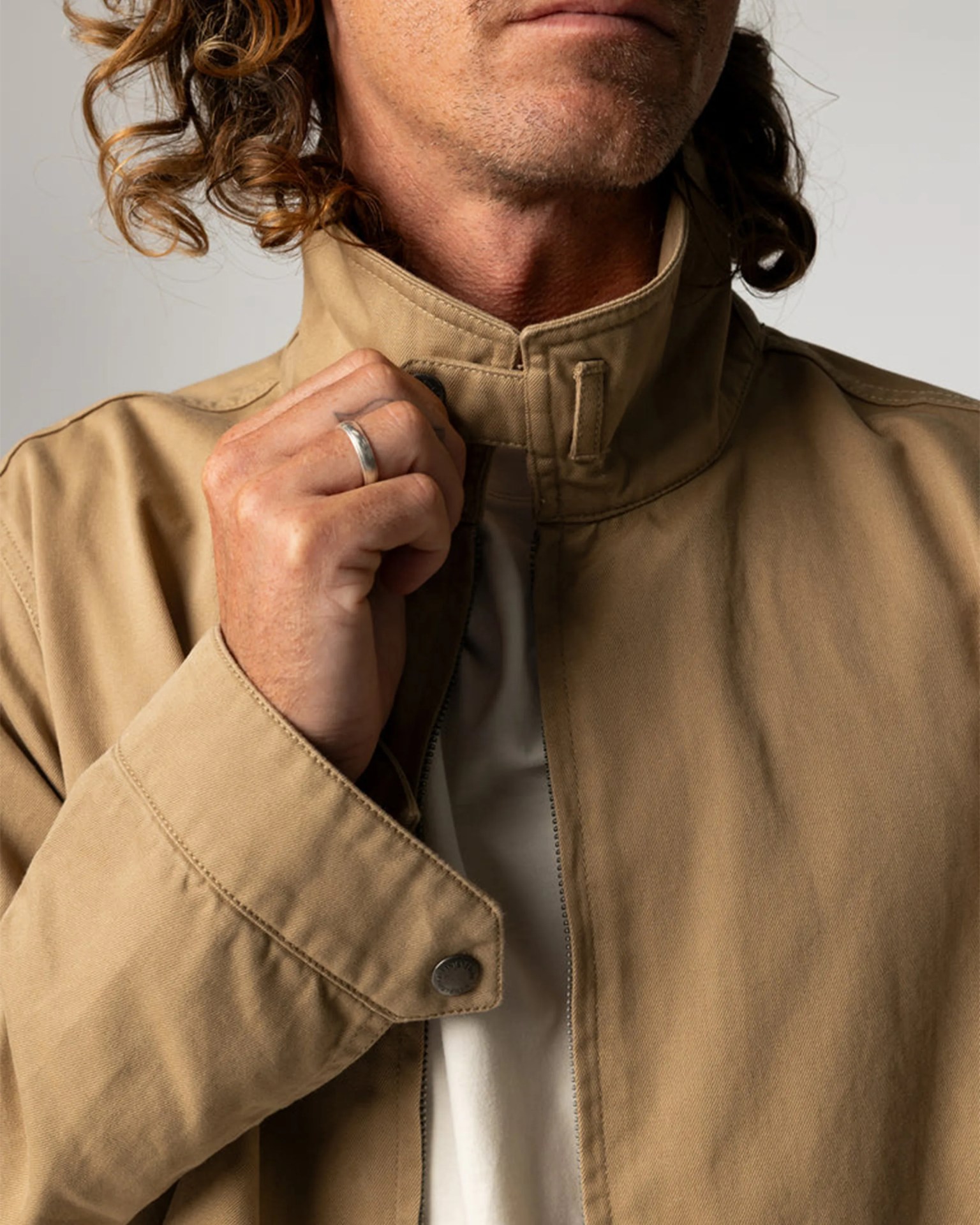 Close-up of a person with curly hair wearing a tan jacket with a high collar and a silver ring on their finger.