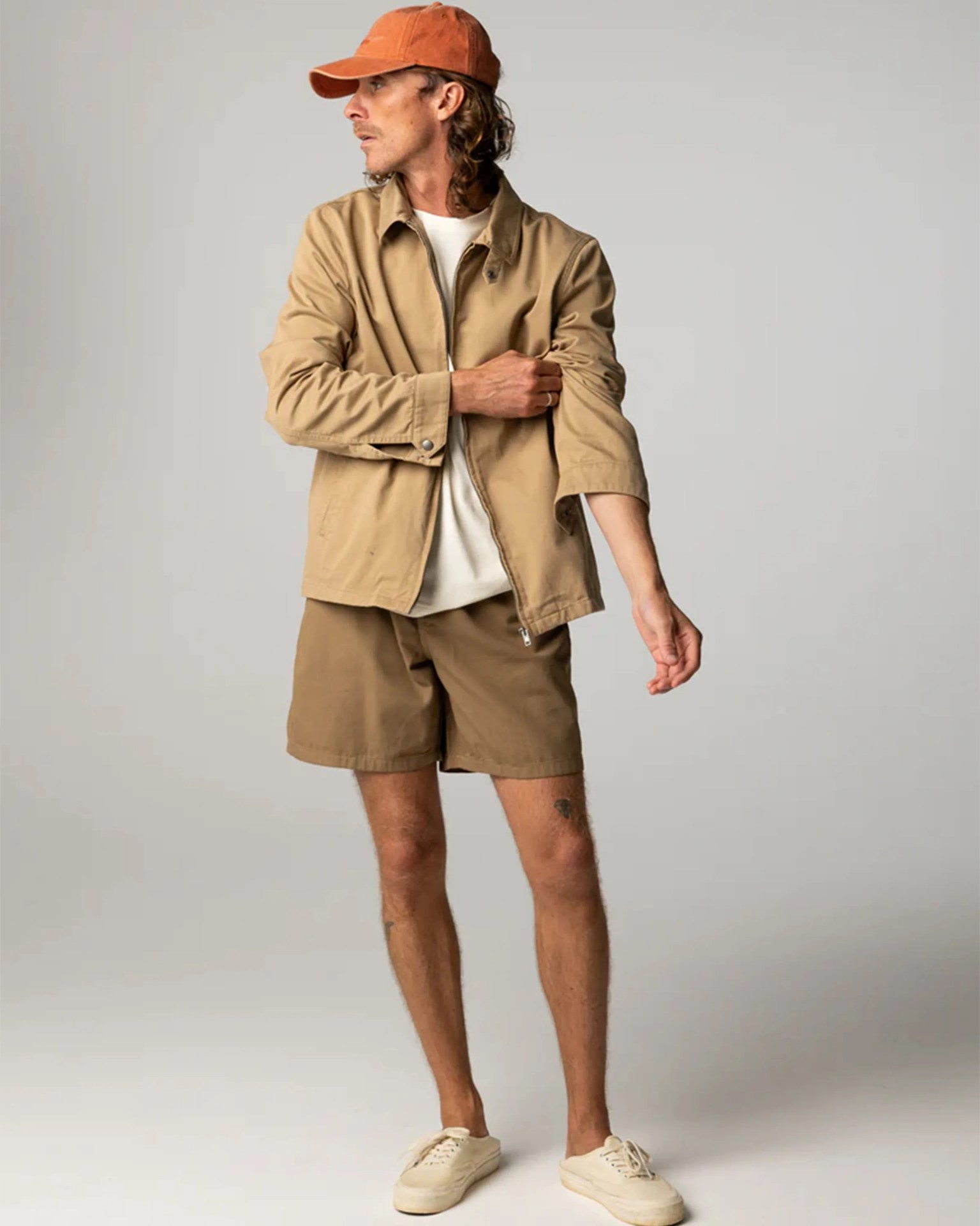 Man wearing a tan jacket, matching shorts, orange cap, and off-white sneakers against a plain background.