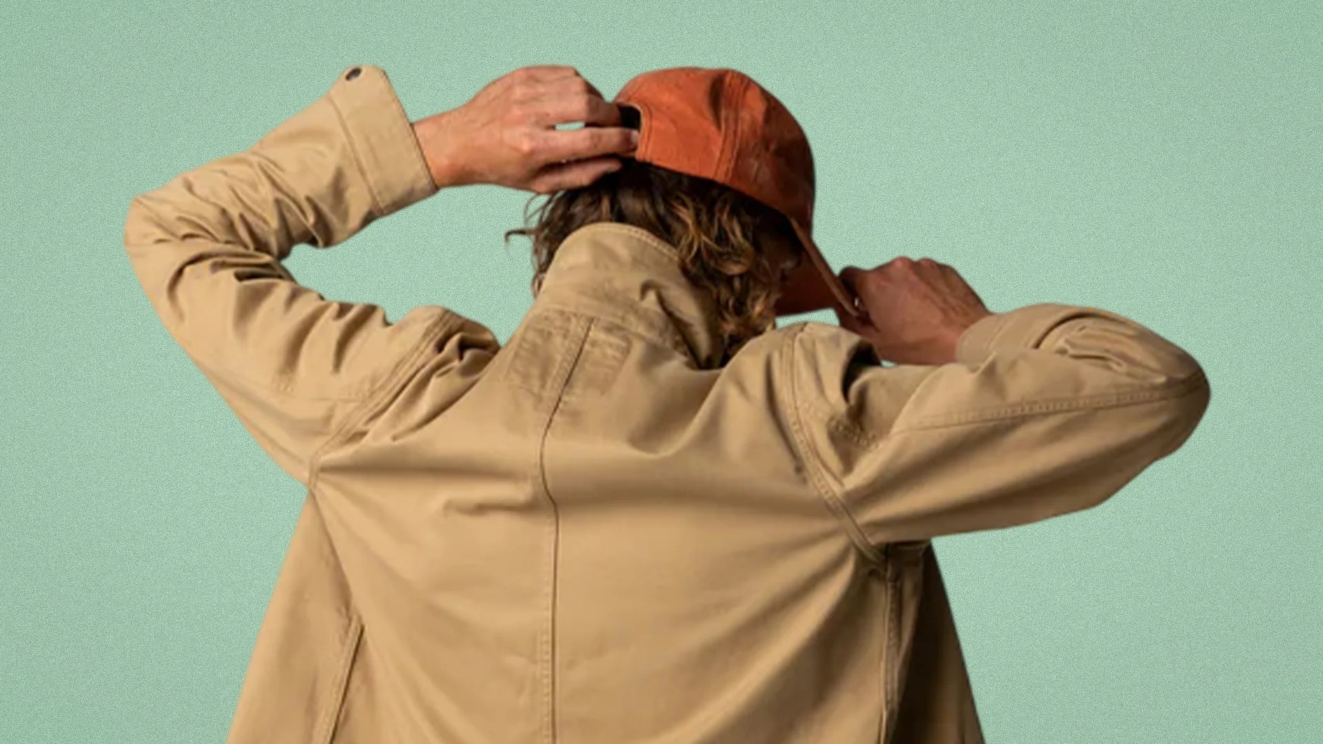 Person wearing a tan jacket adjusting an orange baseball cap against a light green background.