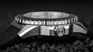 Close-up of a stainless steel watch case with a textured black bezel and a crown featuring an engraved logo, on a black strap.