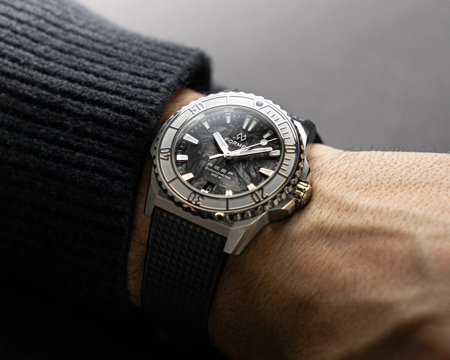 Silver dive watch with black textured dial and black rubber strap worn on a wrist with a black knit sleeve.