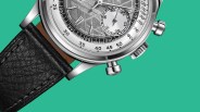 Close-up of a silver chronograph watch with a textured dial, black leather strap, and two push buttons on a teal background.