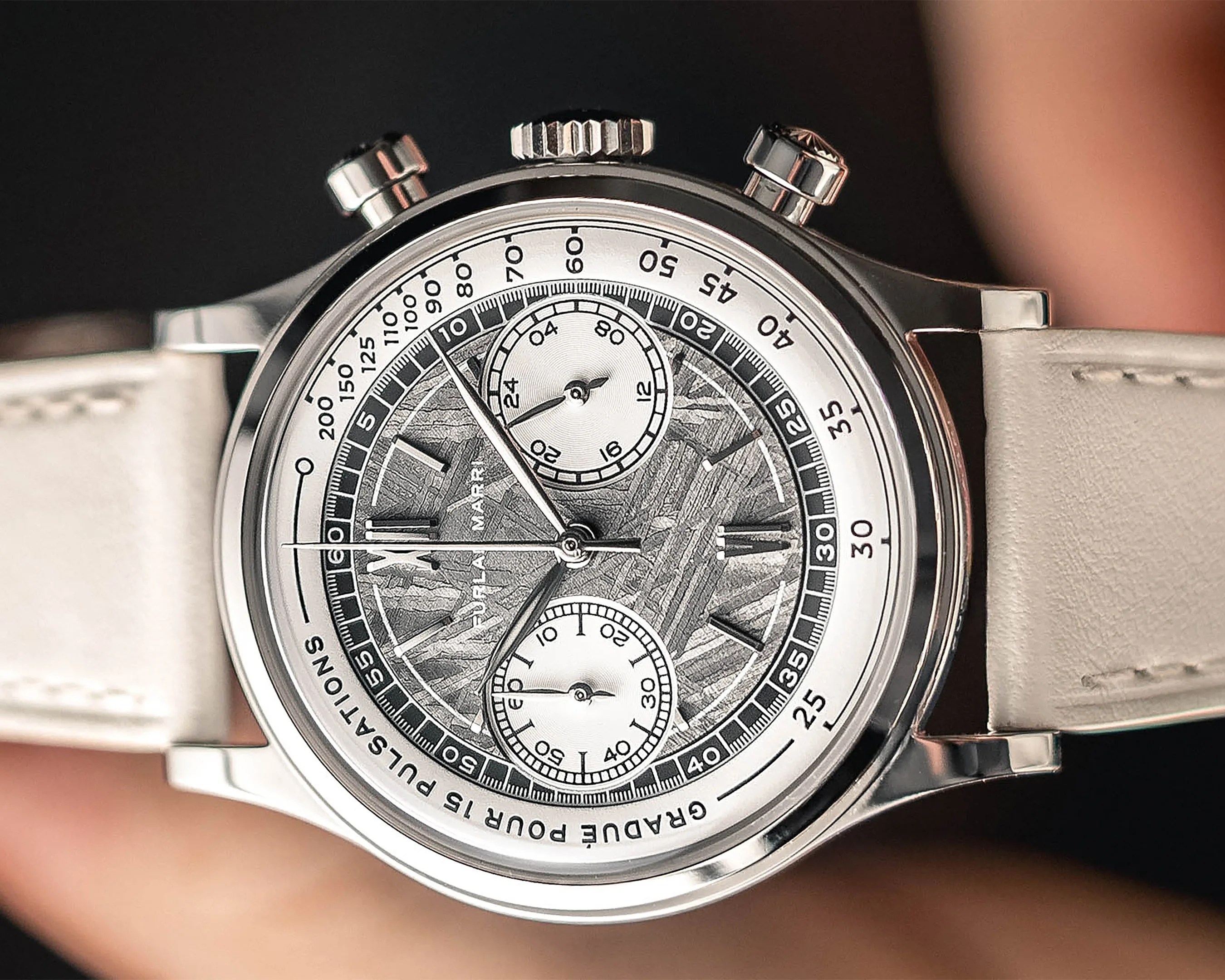 Silver chronograph wristwatch with textured gray dial and white leather strap held in a hand.
