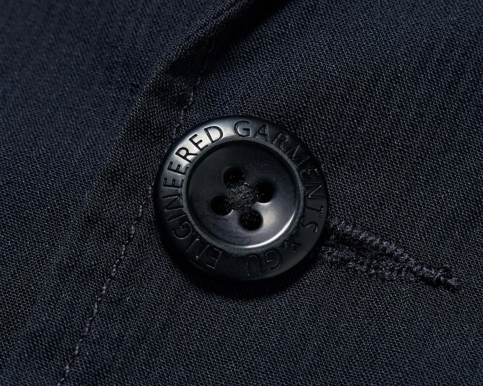 Close-up of a black button on dark fabric with the text "ENGINEERED GARMENTS × GU" embossed around the edge.