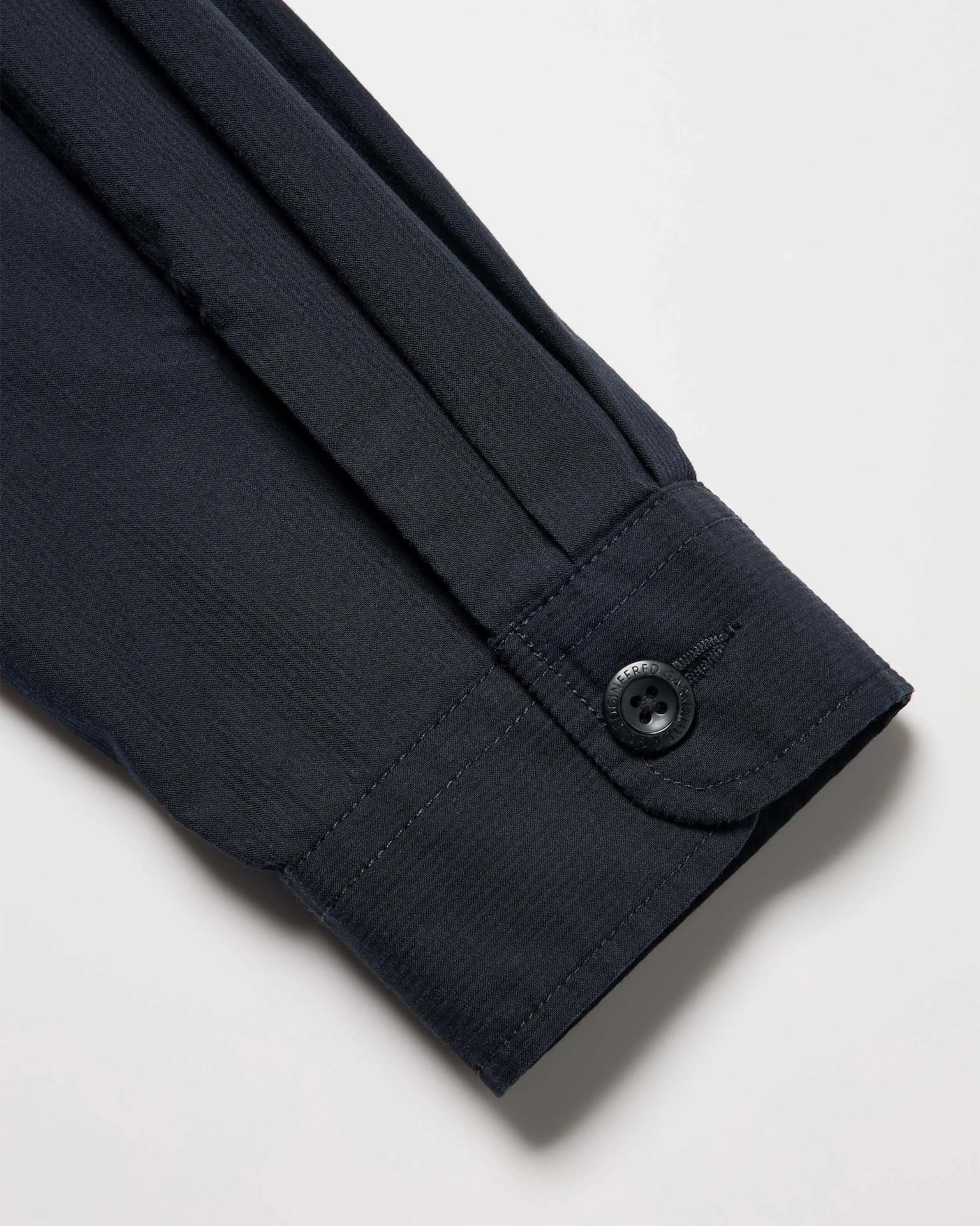 Close-up of a black shirt sleeve cuff with a black button and visible stitching on a light background.