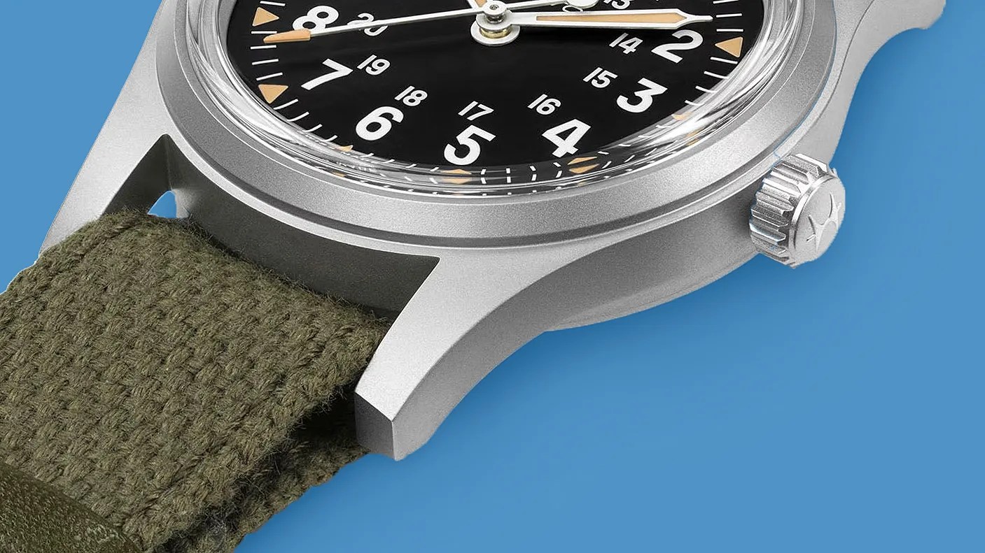 Close-up of a silver watch case with a black dial, beige markers, and an olive green fabric strap on a blue background.