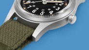 Close-up of a silver watch case with a black dial, beige markers, and an olive green fabric strap on a blue background.