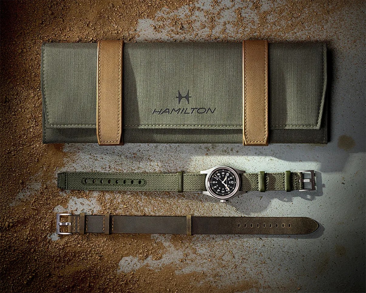 Green fabric Hamilton watch roll with tan leather straps, green fabric strap watch with black dial, and brown leather strap on a textured surface.