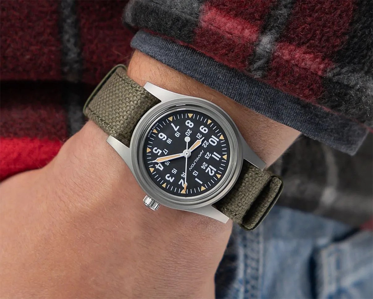 Wristwatch with black dial, white numerals, and olive green fabric strap worn on a wrist.