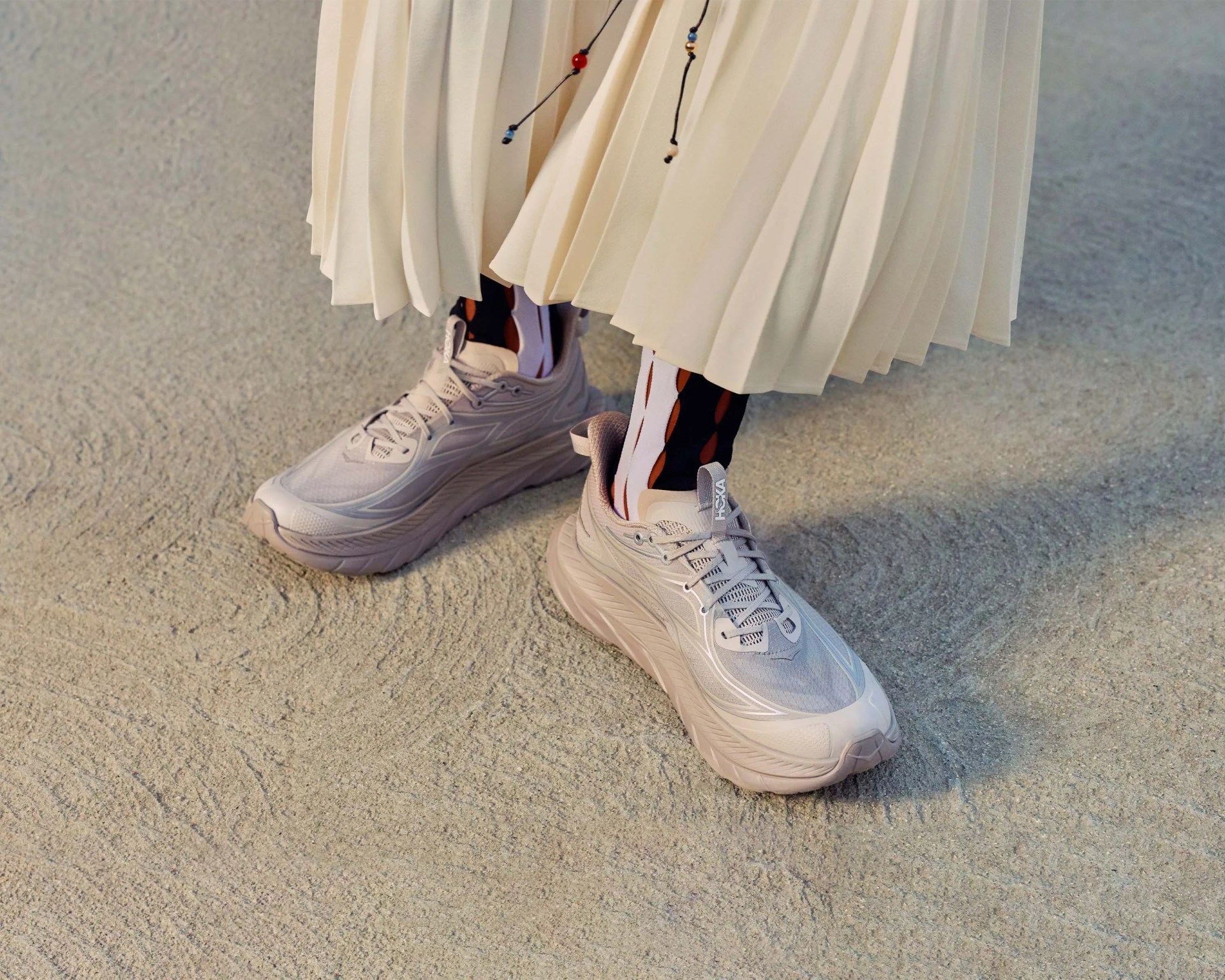 Light gray Hoka running shoes worn with patterned socks and a cream pleated skirt on textured ground.