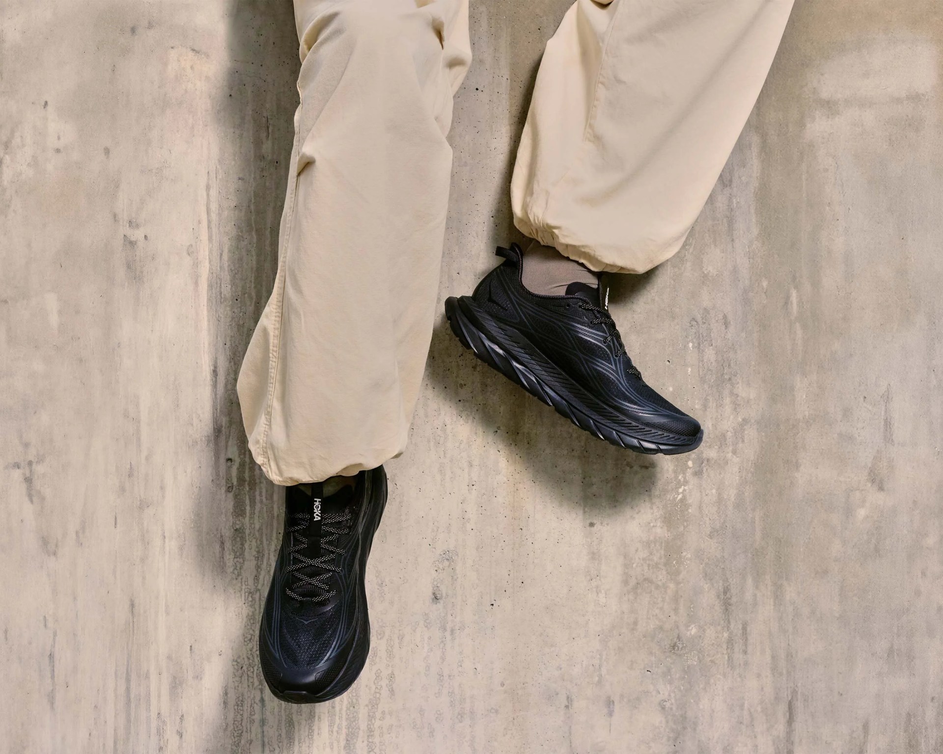 Pair of black Hoka running shoes worn with beige pants against a concrete wall.