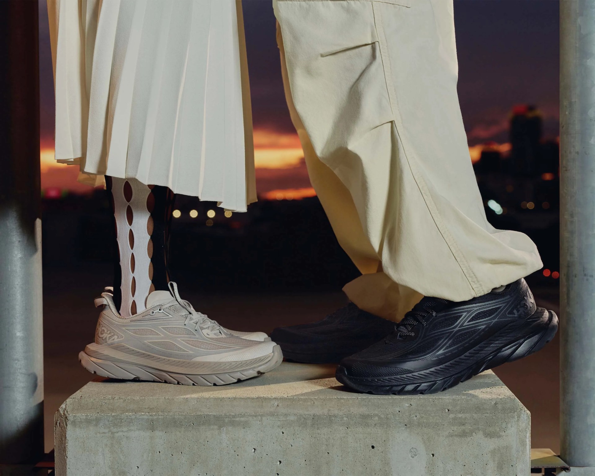 Pair of light beige and black running shoes worn by two people standing on a concrete block at sunset.