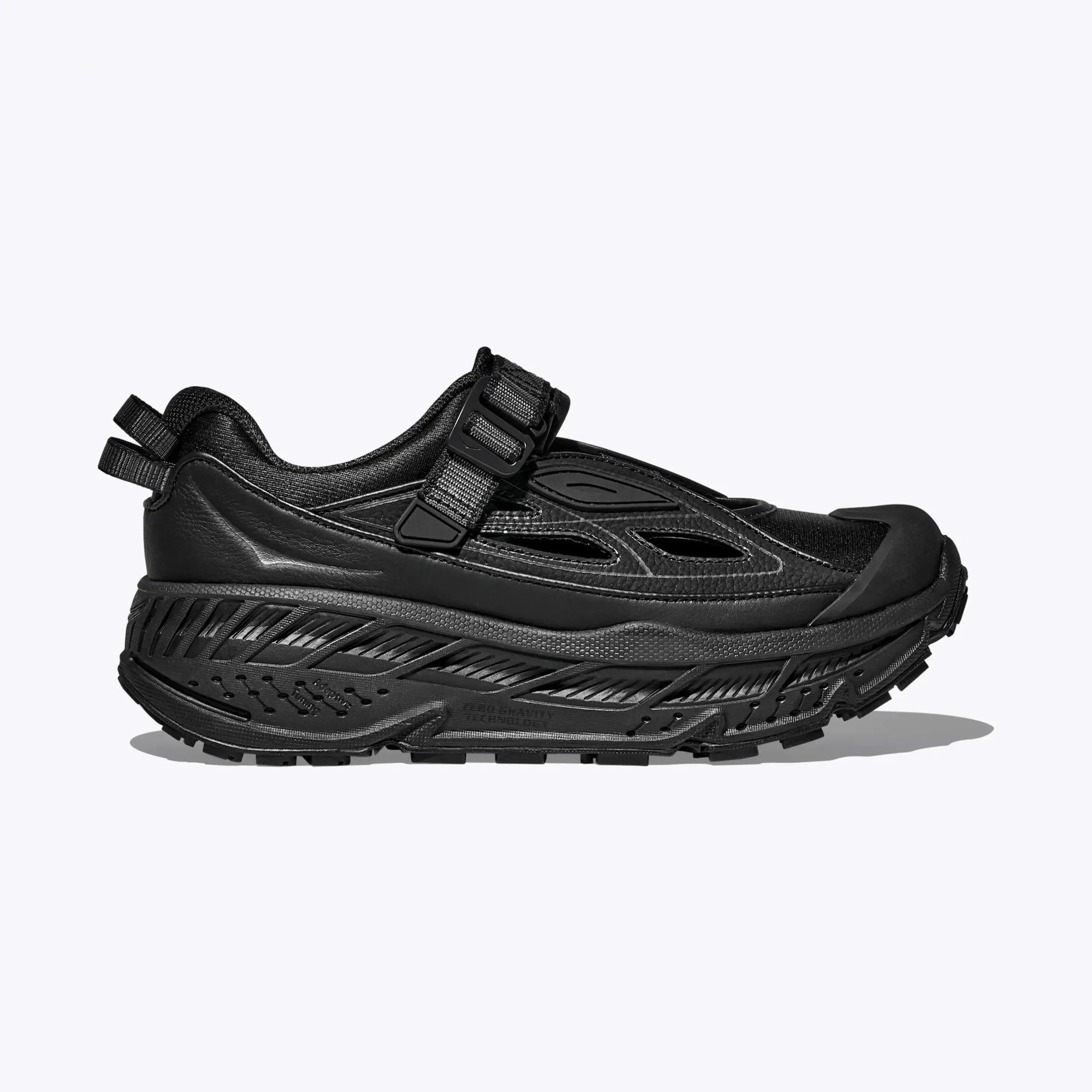 All-black athletic shoe with thick textured sole, mesh and leather upper, and adjustable strap closure.
