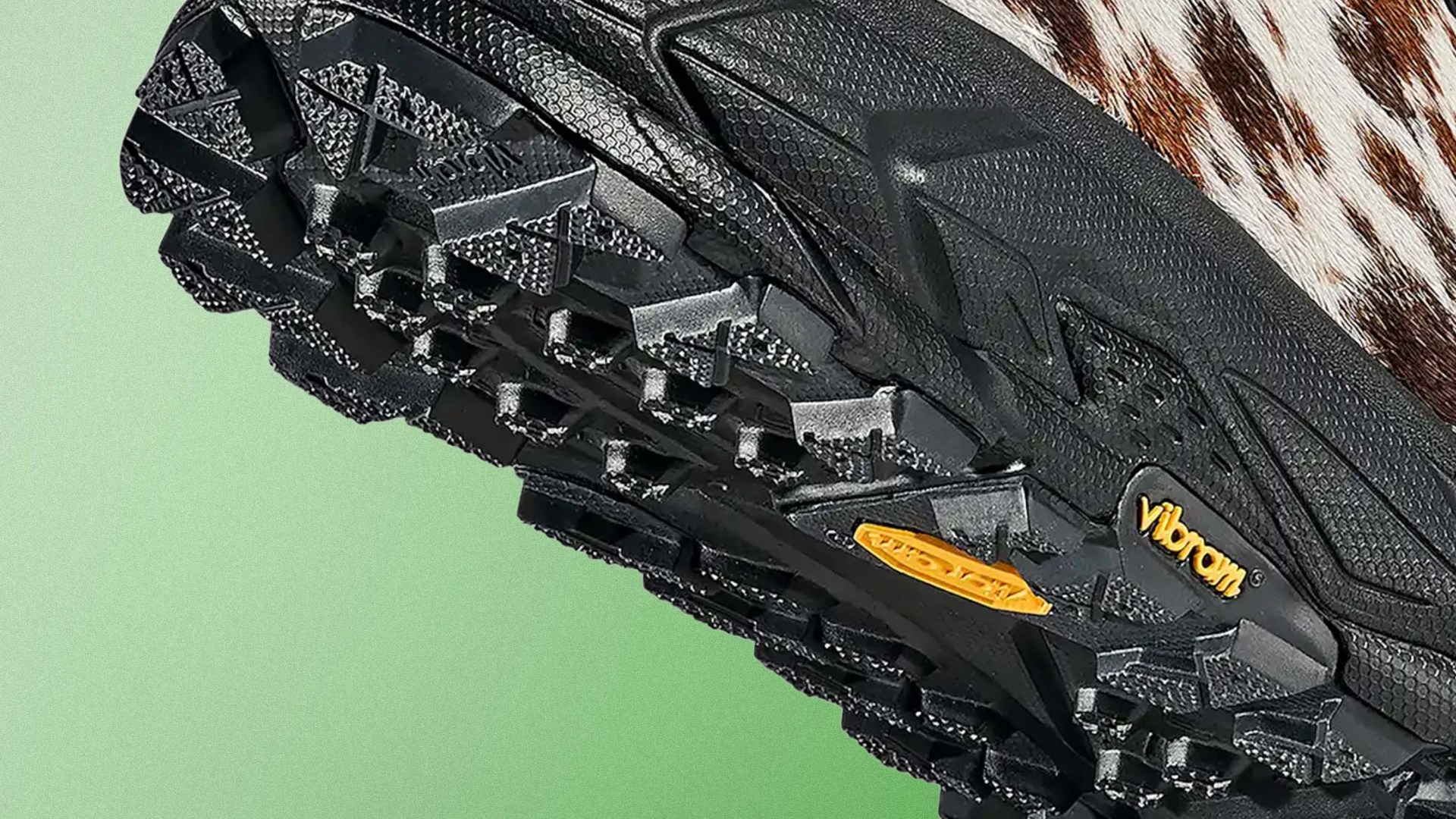 Close-up of a black Vibram shoe sole with deep treads and yellow Vibram logo on a green background.