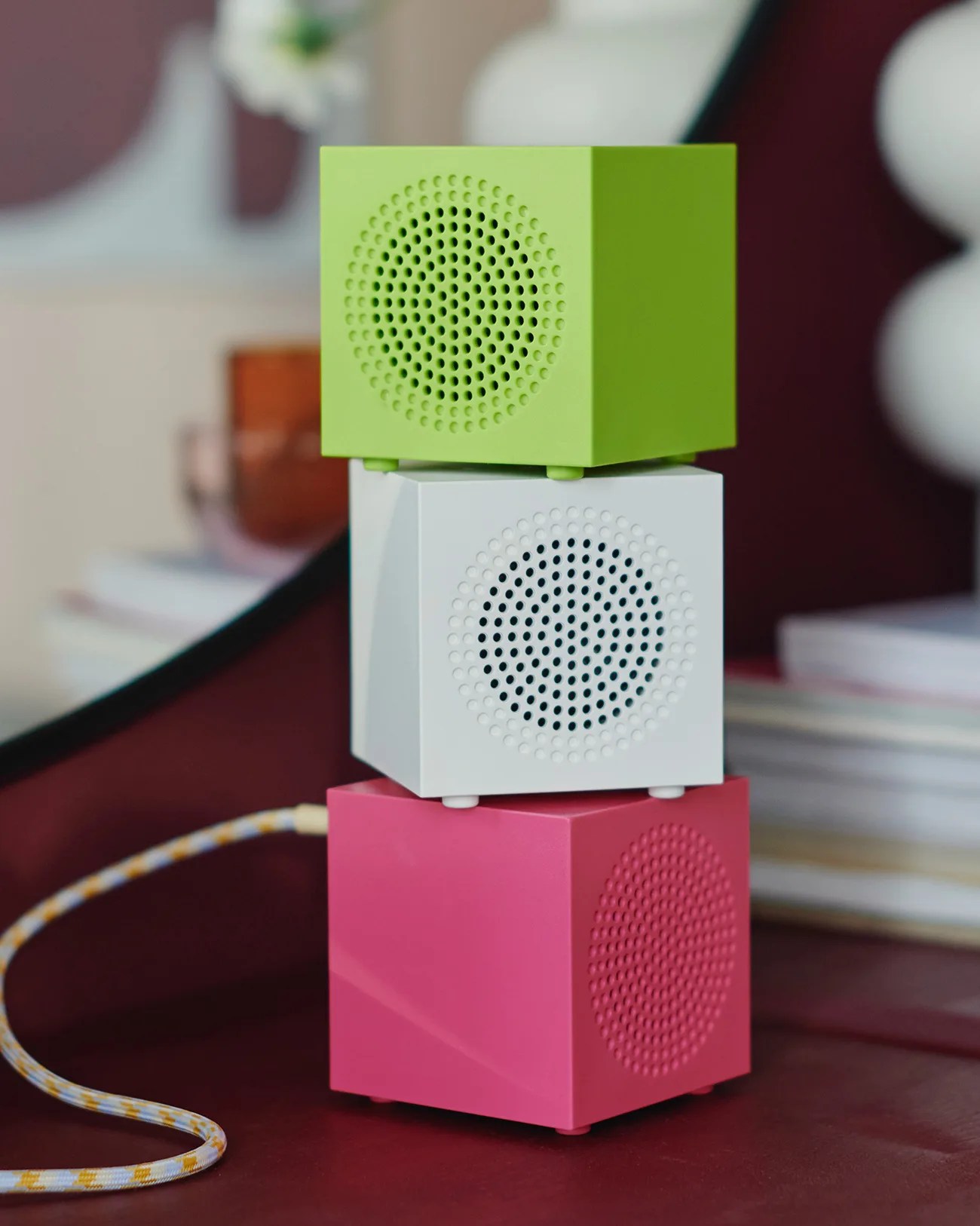 Stack of three small cube-shaped speakers in green, white, and pink on a red surface with a braided cable attached.