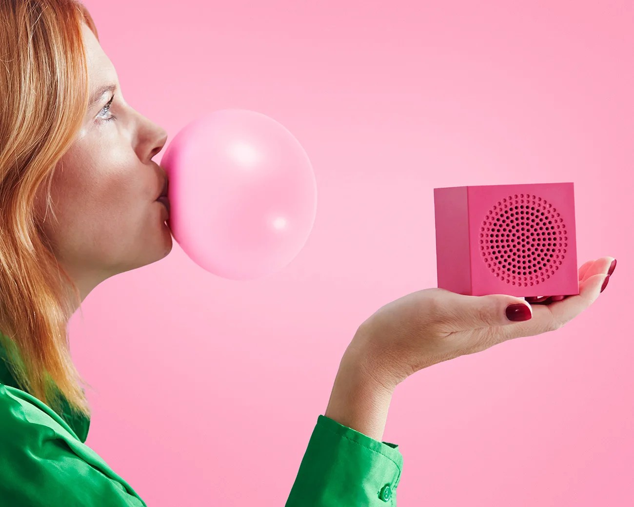 Person in green shirt blowing a pink bubblegum bubble while holding a small pink speaker in a hand against a pink background.