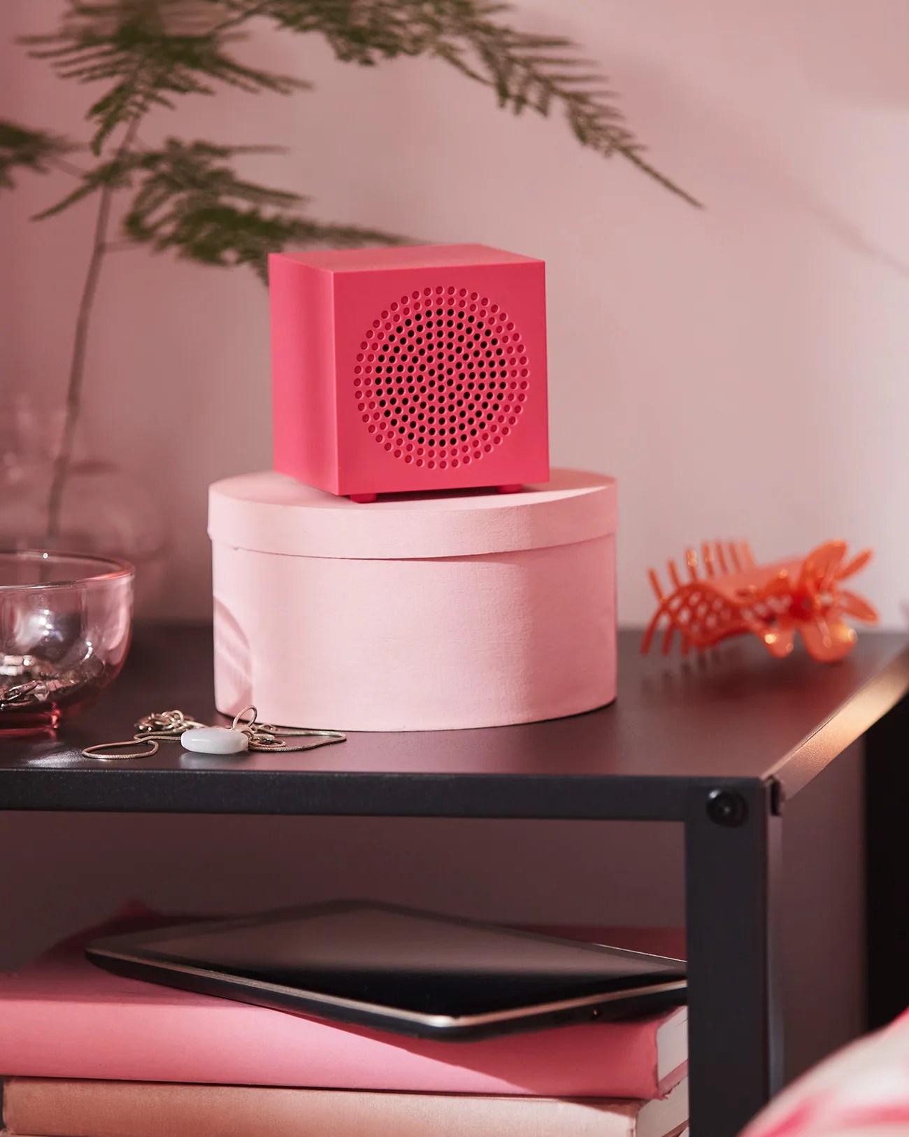 Pink cube-shaped speaker with circular perforated front on a round pink box on a black shelf.