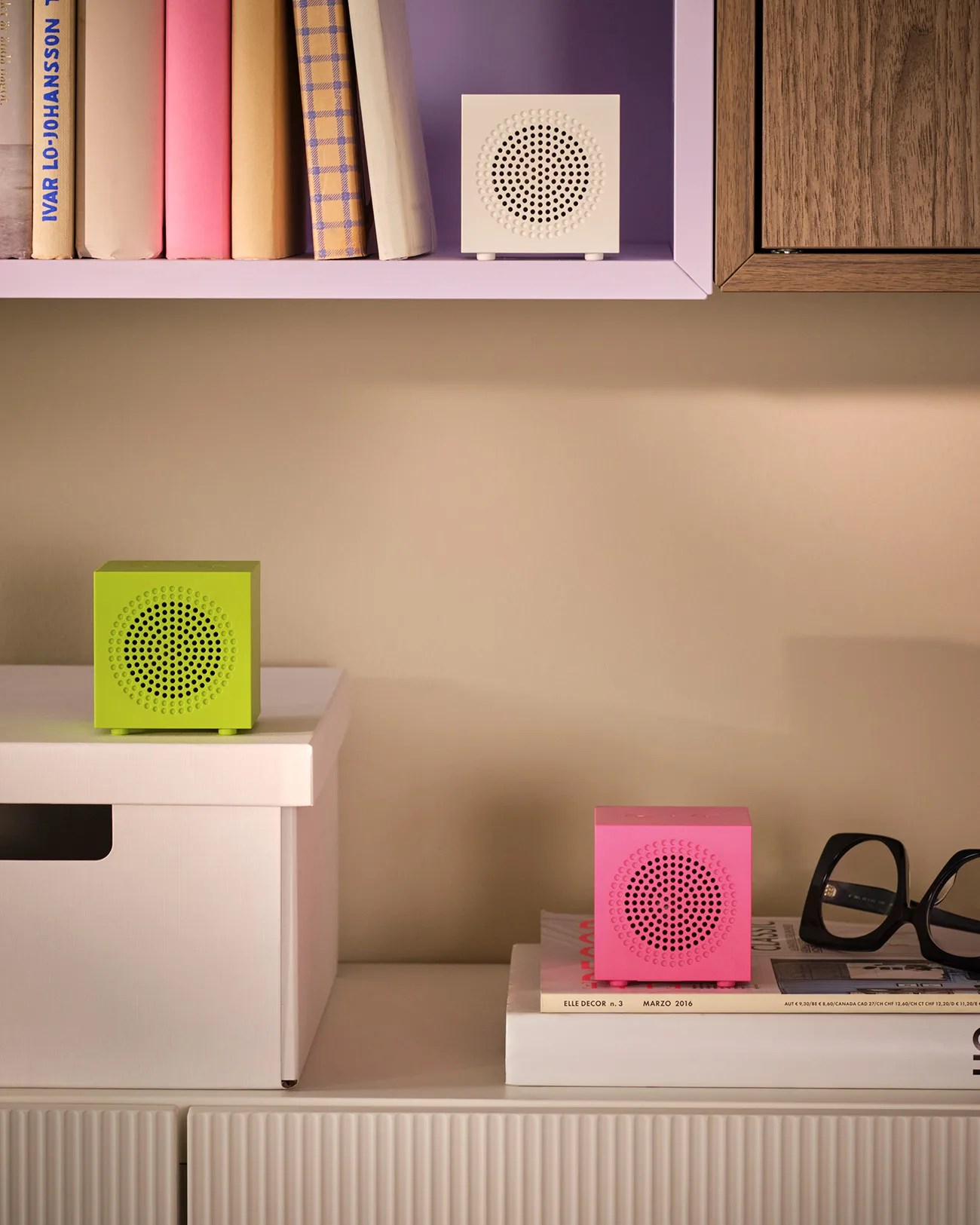 Three small cube-shaped speakers in white, lime green, and pink placed on shelves and books in a room.