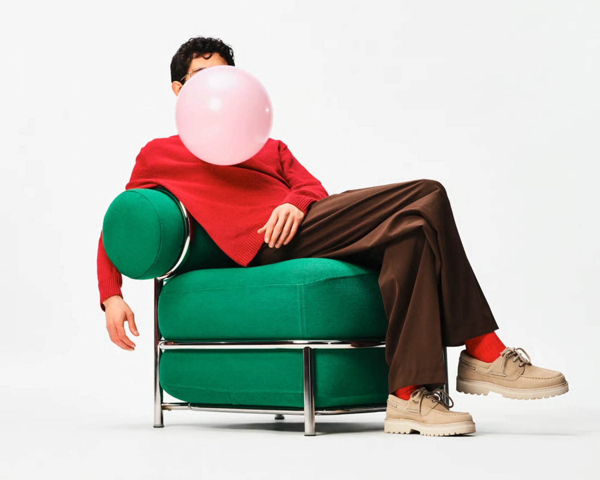 Person in red sweater and brown pants blowing a pink bubblegum bubble while reclining on a green cushioned chair with metal frame.
