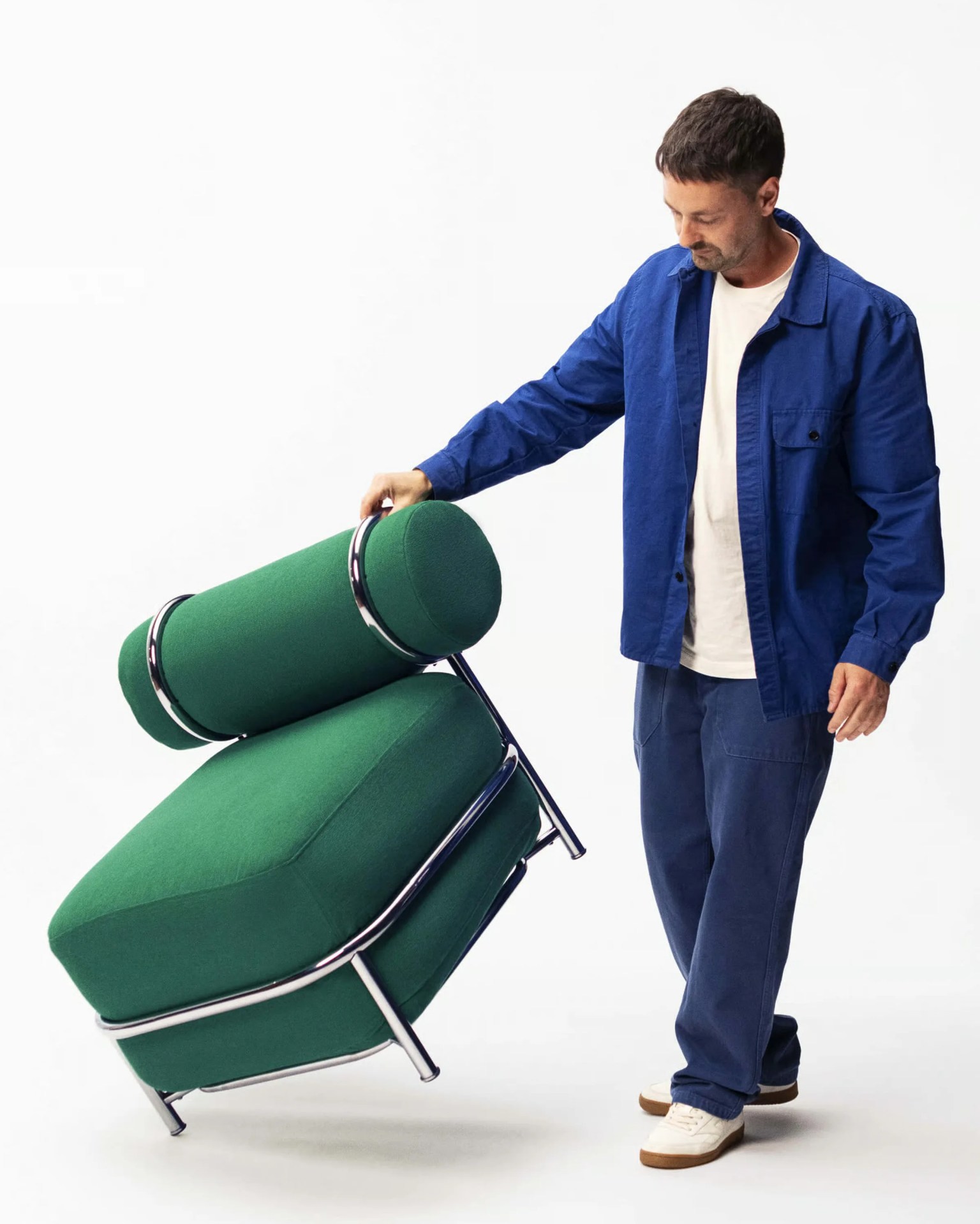 Man in blue jacket and pants holding a green upholstered chair with chrome frame tilted on one leg.