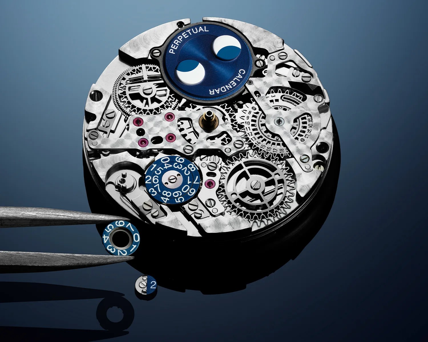 Close-up of a silver watch movement with blue calendar and moon phase dials, held by tweezers on a dark reflective surface.