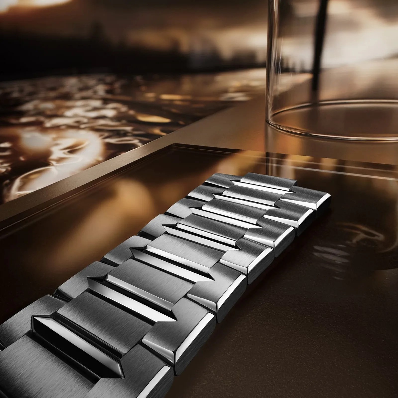 Brushed and polished stainless steel watch bracelet links on a reflective bronze surface next to a glass.