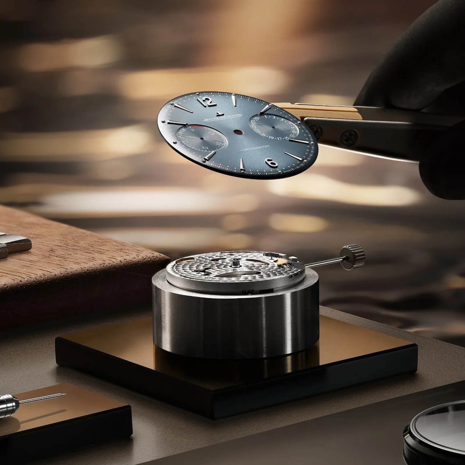 Close-up of a watch dial held by tweezers above a mechanical watch movement on a workbench.