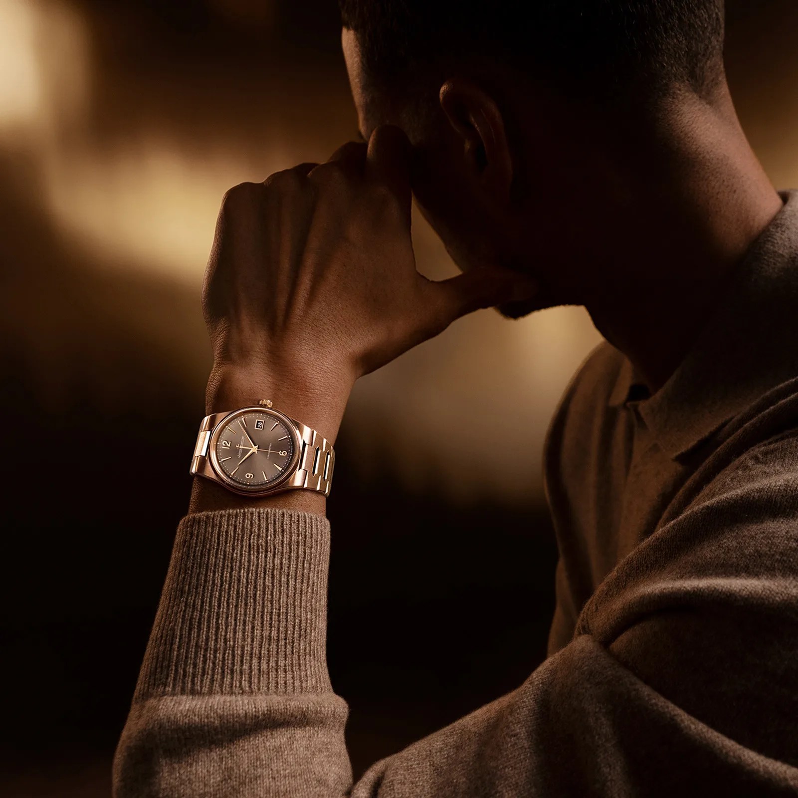 Close-up of a wrist wearing a gold metal bracelet watch with a dark dial and date display, worn with a brown sweater.