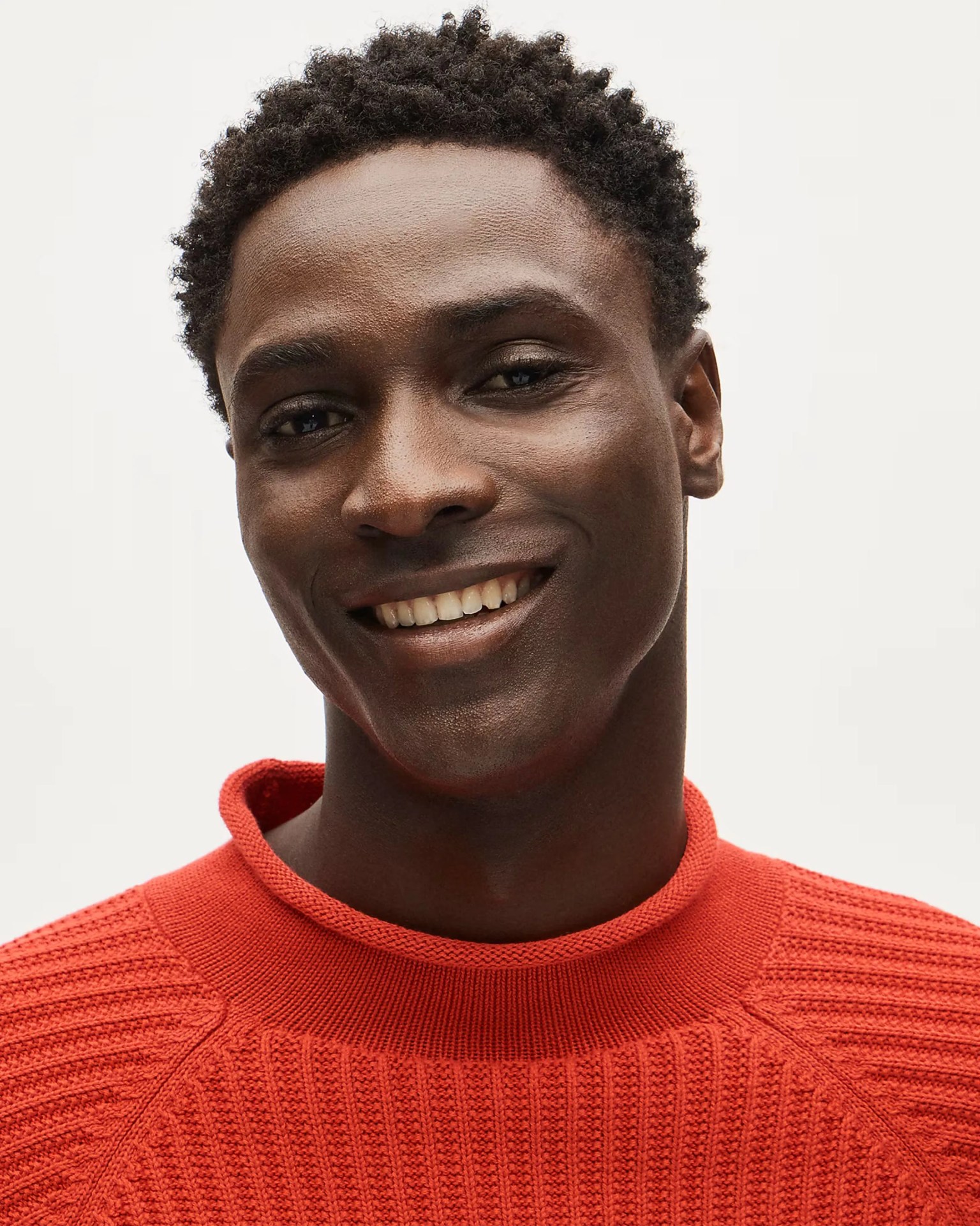 Close-up of a person smiling, wearing a textured bright orange knit sweater with a round neckline.