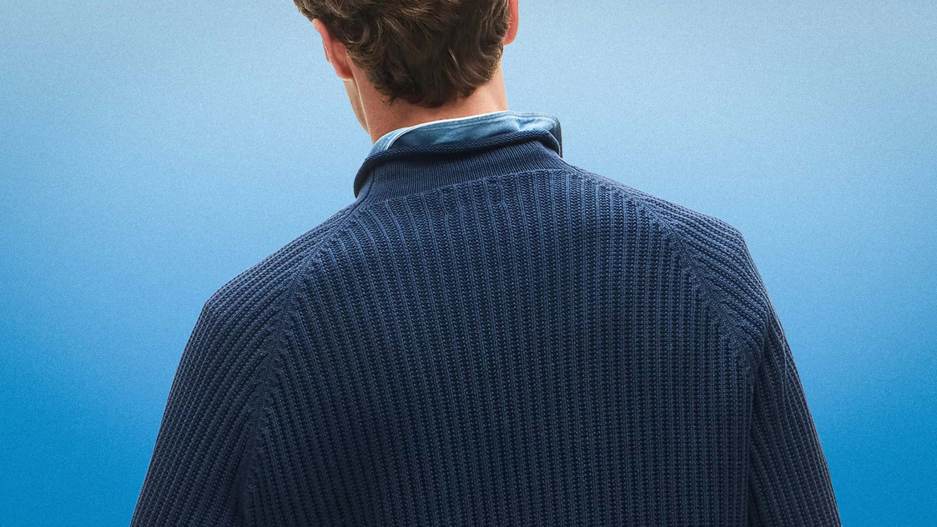 Man wearing a navy blue textured knit sweater over a light blue collared shirt, viewed from the back.
