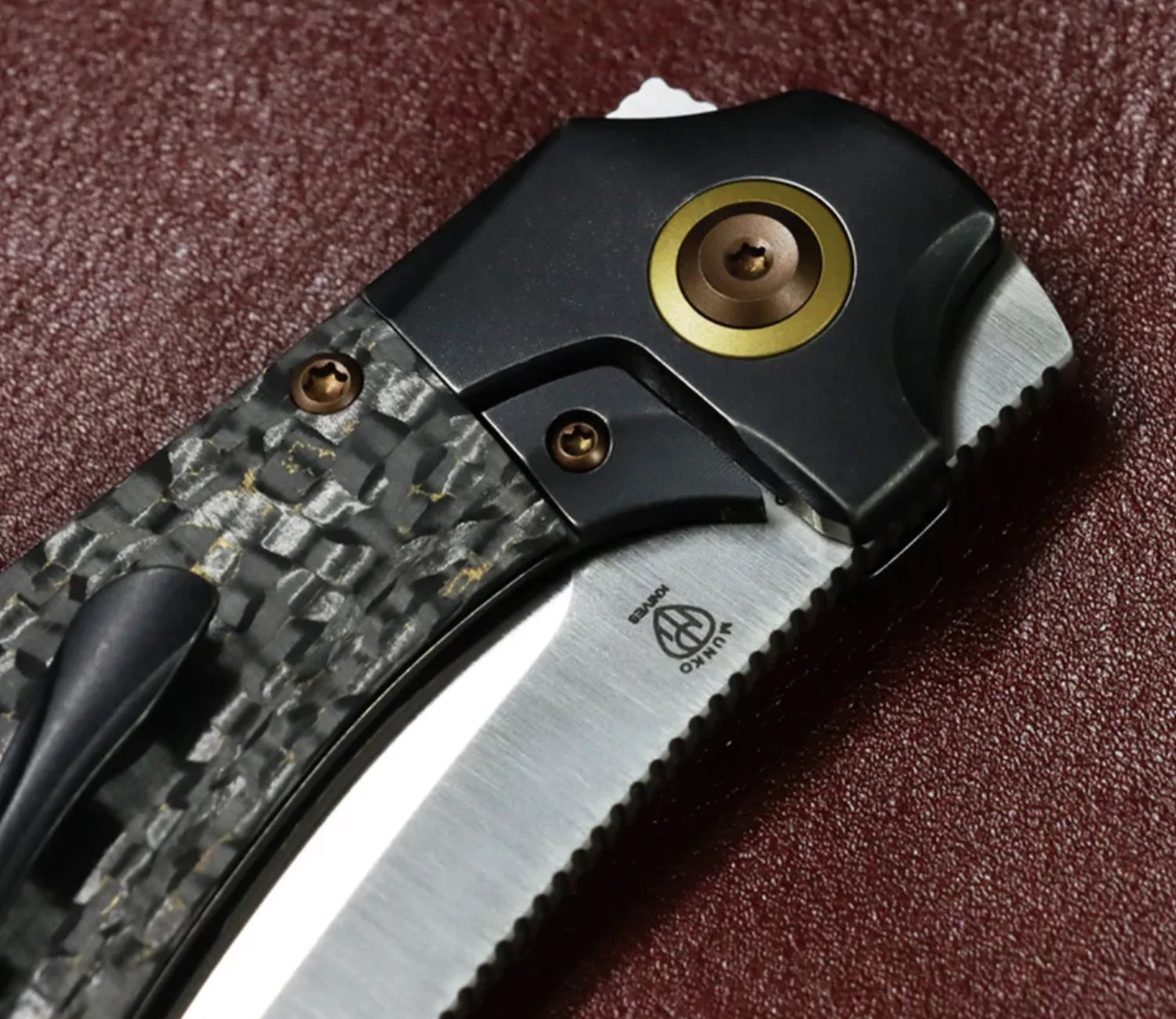 Close-up of a folding knife with a carbon fiber handle, black and gold pivot, and partially serrated stainless steel blade.