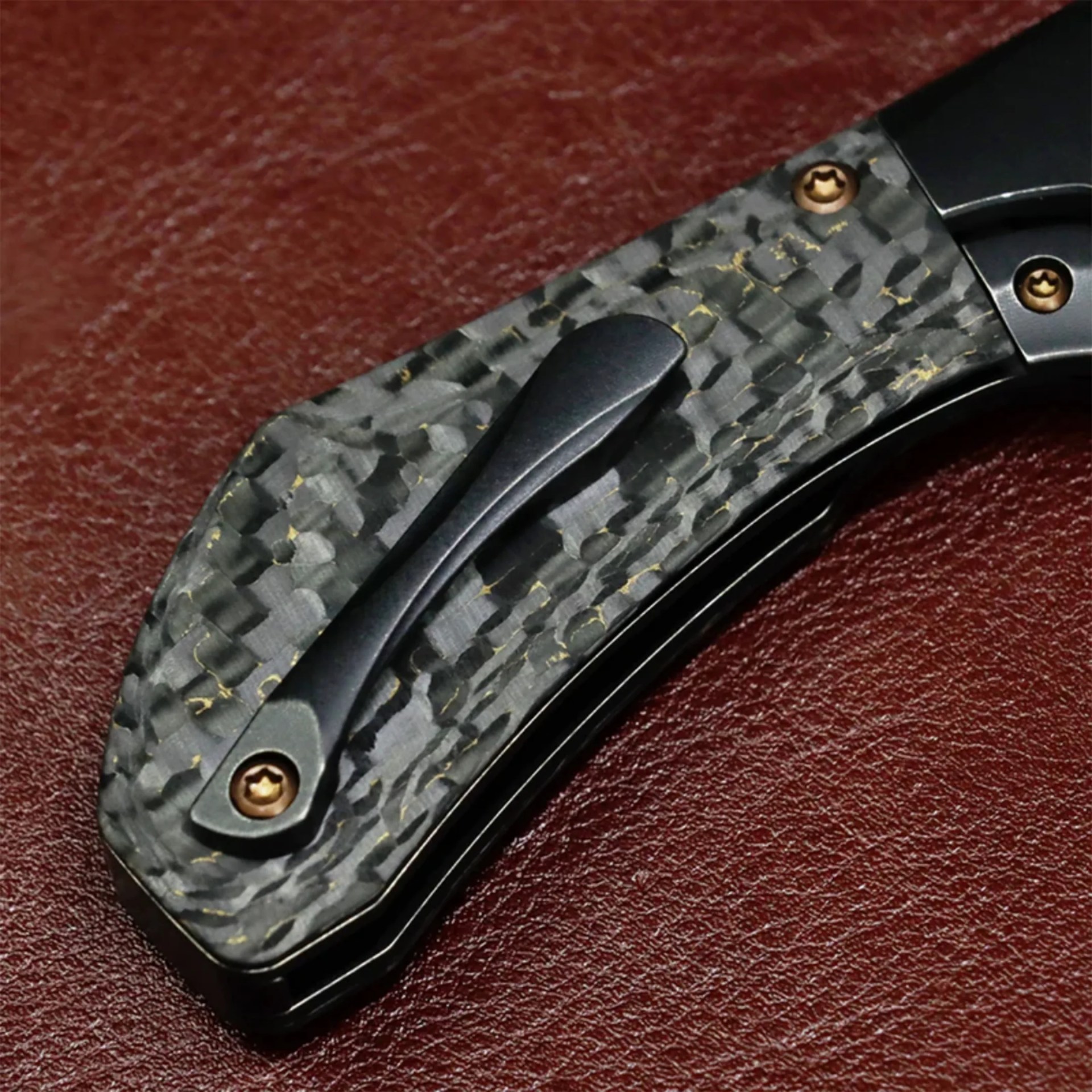 Close-up of a black and gray carbon fiber knife handle with gold screws on a textured brown surface.