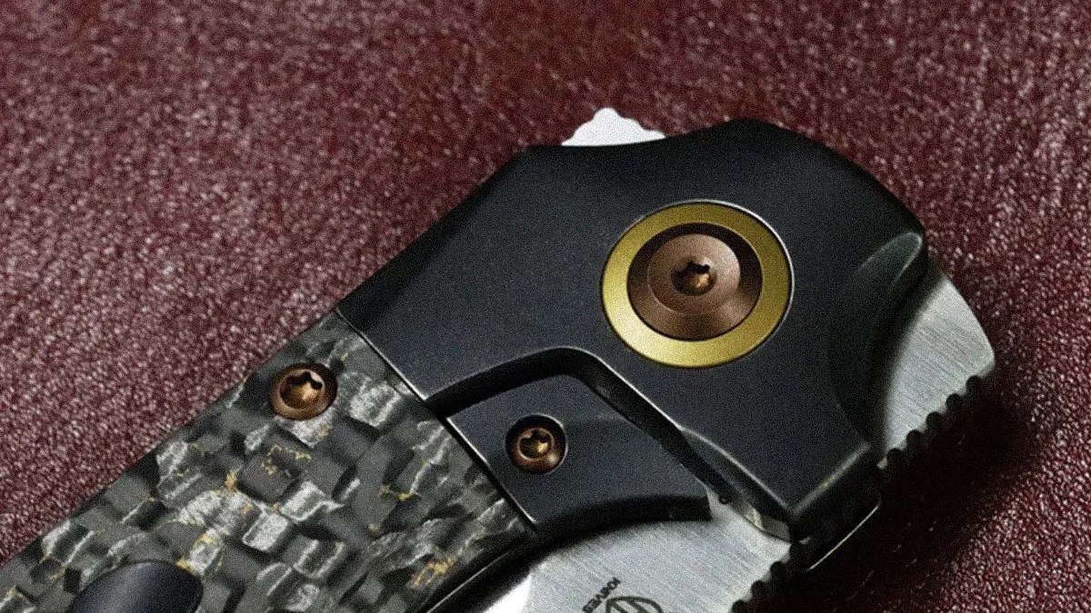Close-up of a folding knife handle with textured carbon fiber and black metal parts with bronze screws.