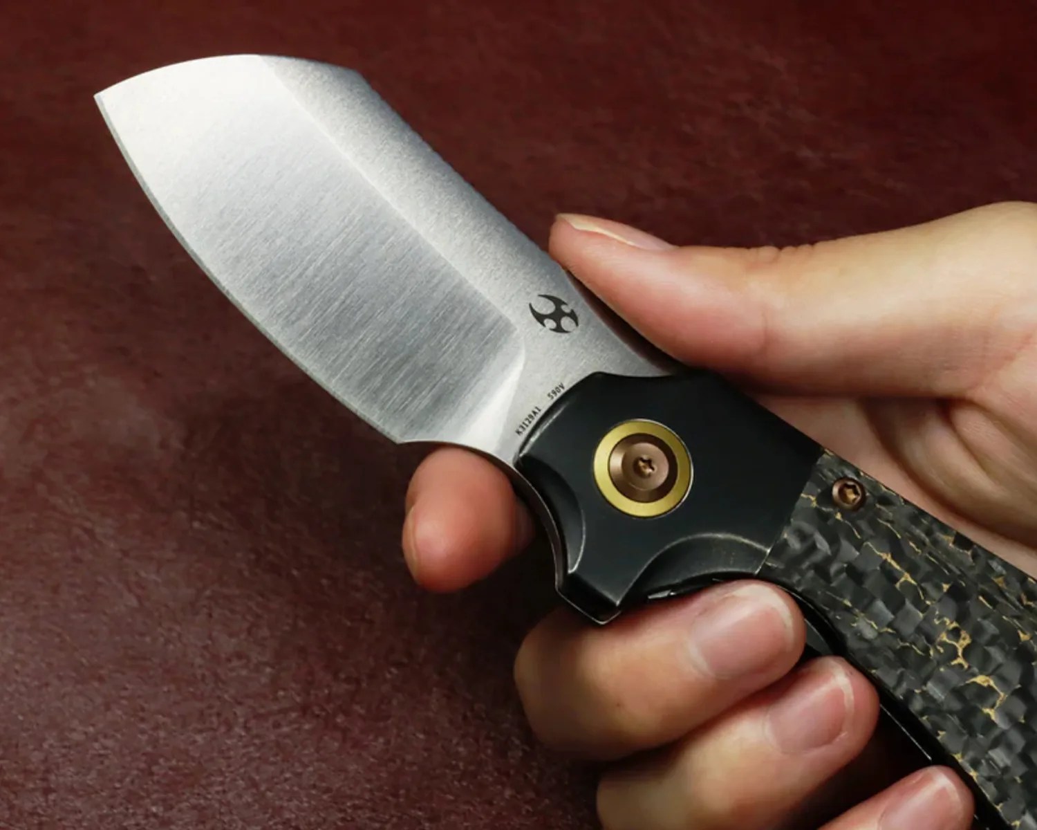 Hand holding a folding knife with a brushed steel blade and a black carbon fiber handle with gold accents.
