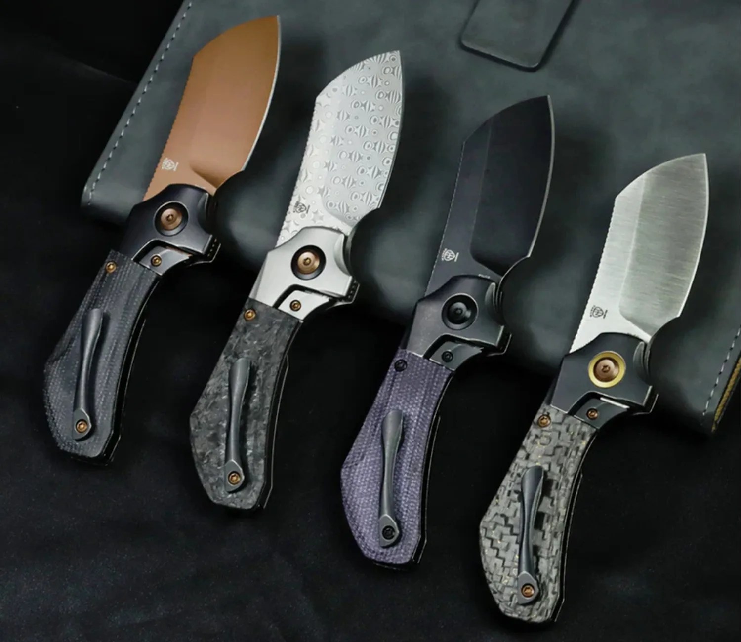 Four folding knives with textured handles and different blade finishes laid on a dark surface.
