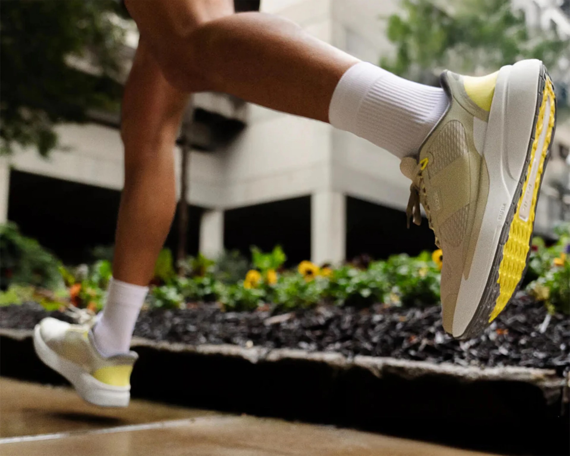 Beige and yellow running shoes with white soles worn with white crew socks outdoors near greenery and a building.