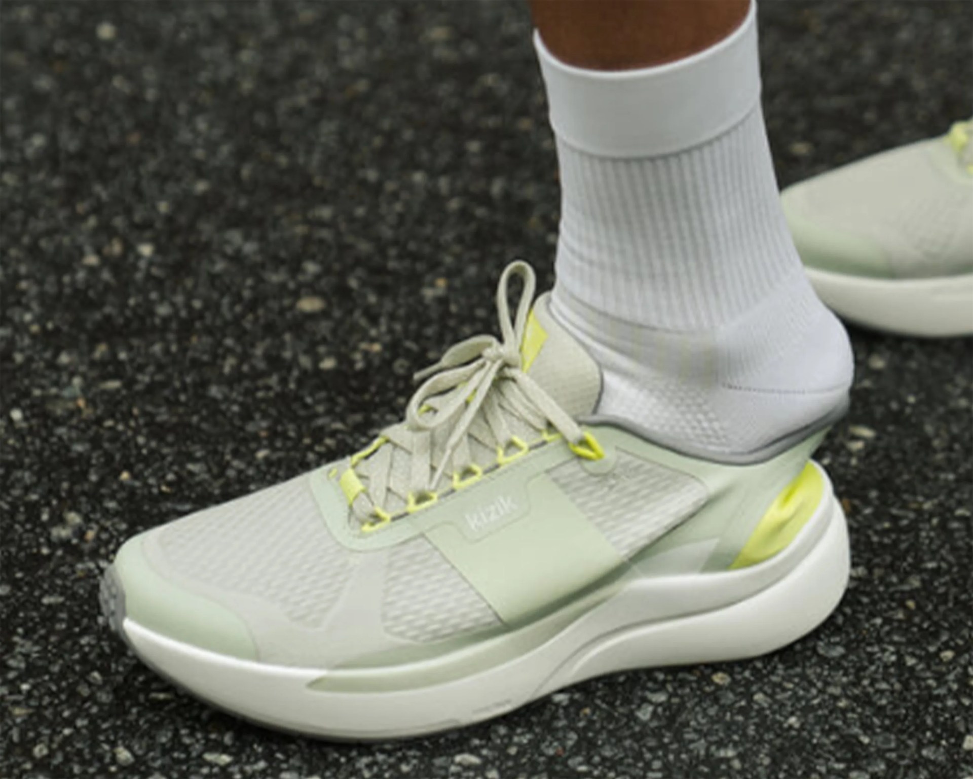 Light green and white athletic shoe with yellow accents worn with white ribbed socks on black asphalt.