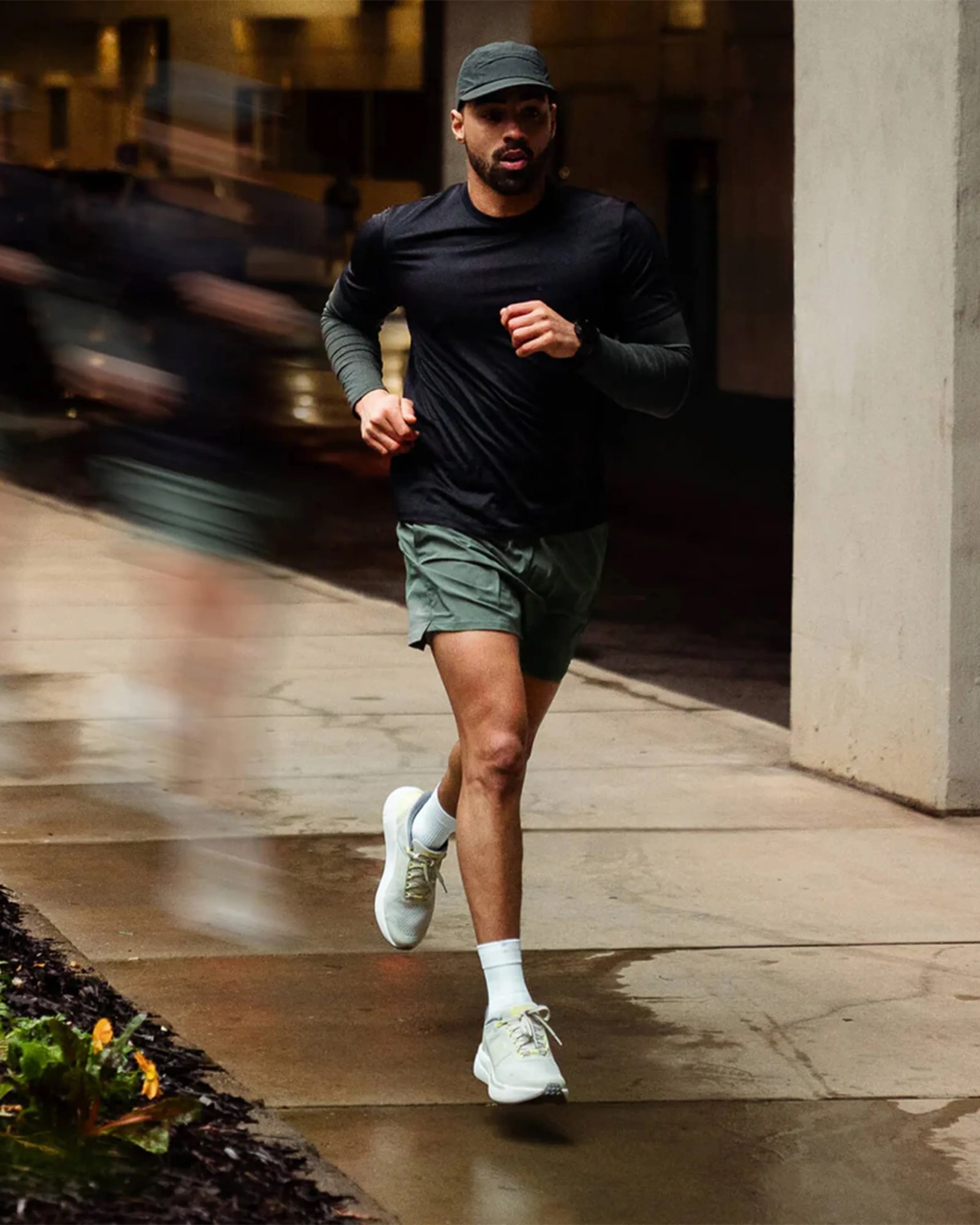 Man running on a wet urban sidewalk wearing a black cap, black long-sleeve shirt, green shorts, white socks, and white running shoes.