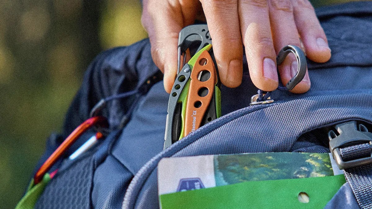 Hand placing a green and orange multitool with carabiner clip into the front pocket of a blue backpack.