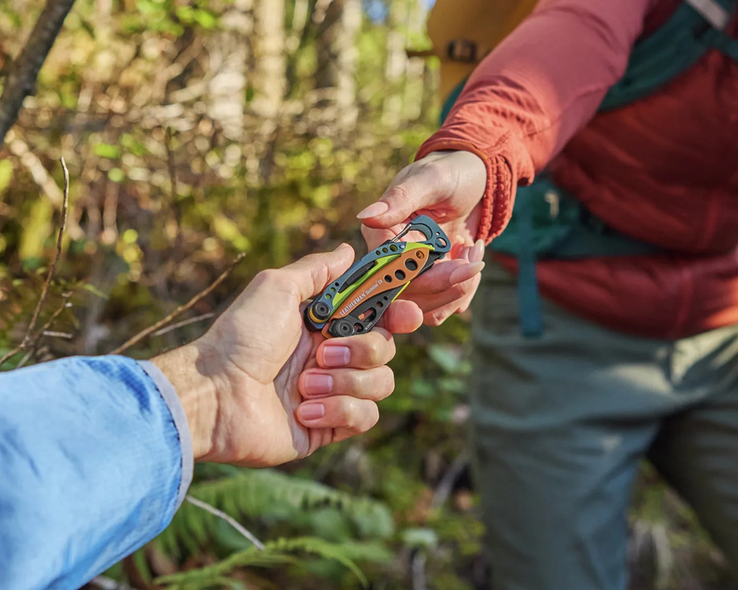 Two hands exchanging a Leatherman multi-tool with orange and green accents in an outdoor forest setting.