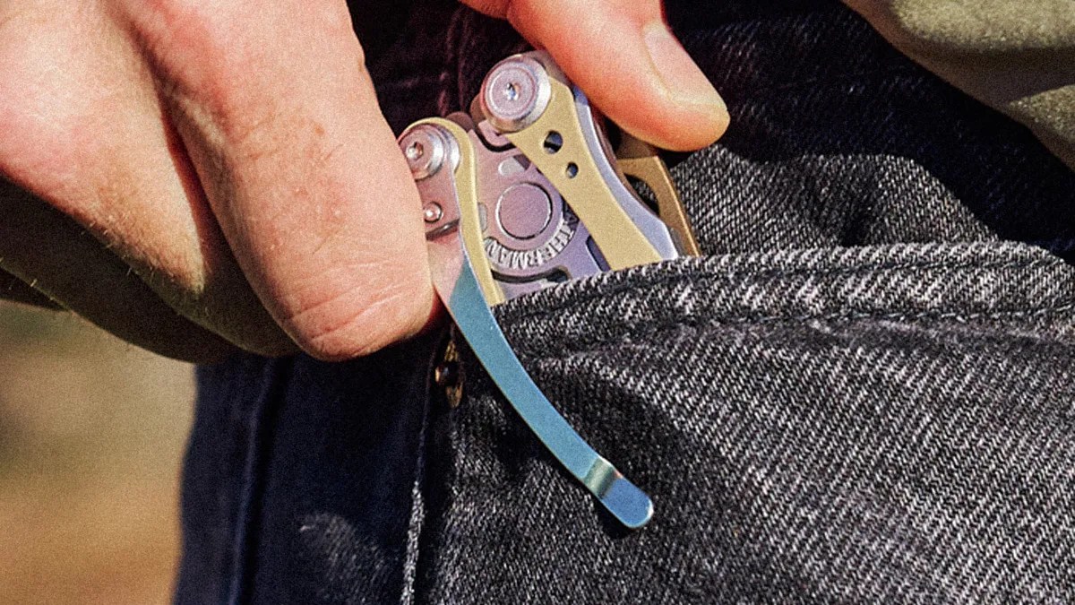 Hand placing a silver and gold multitool with a clip into a dark denim pocket.