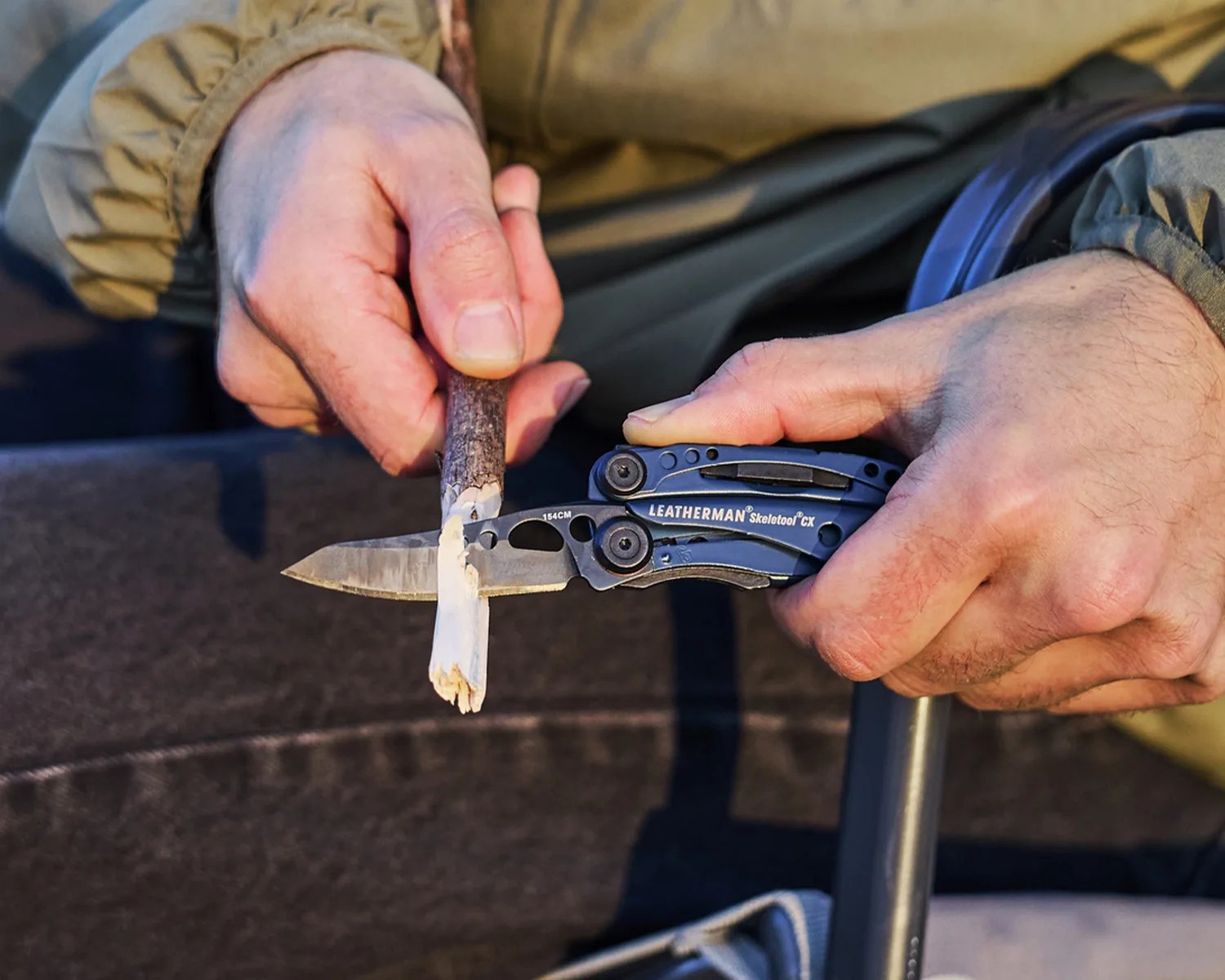 Hands holding a Leatherman Skeletool CX multitool knife carving a small wooden stick.