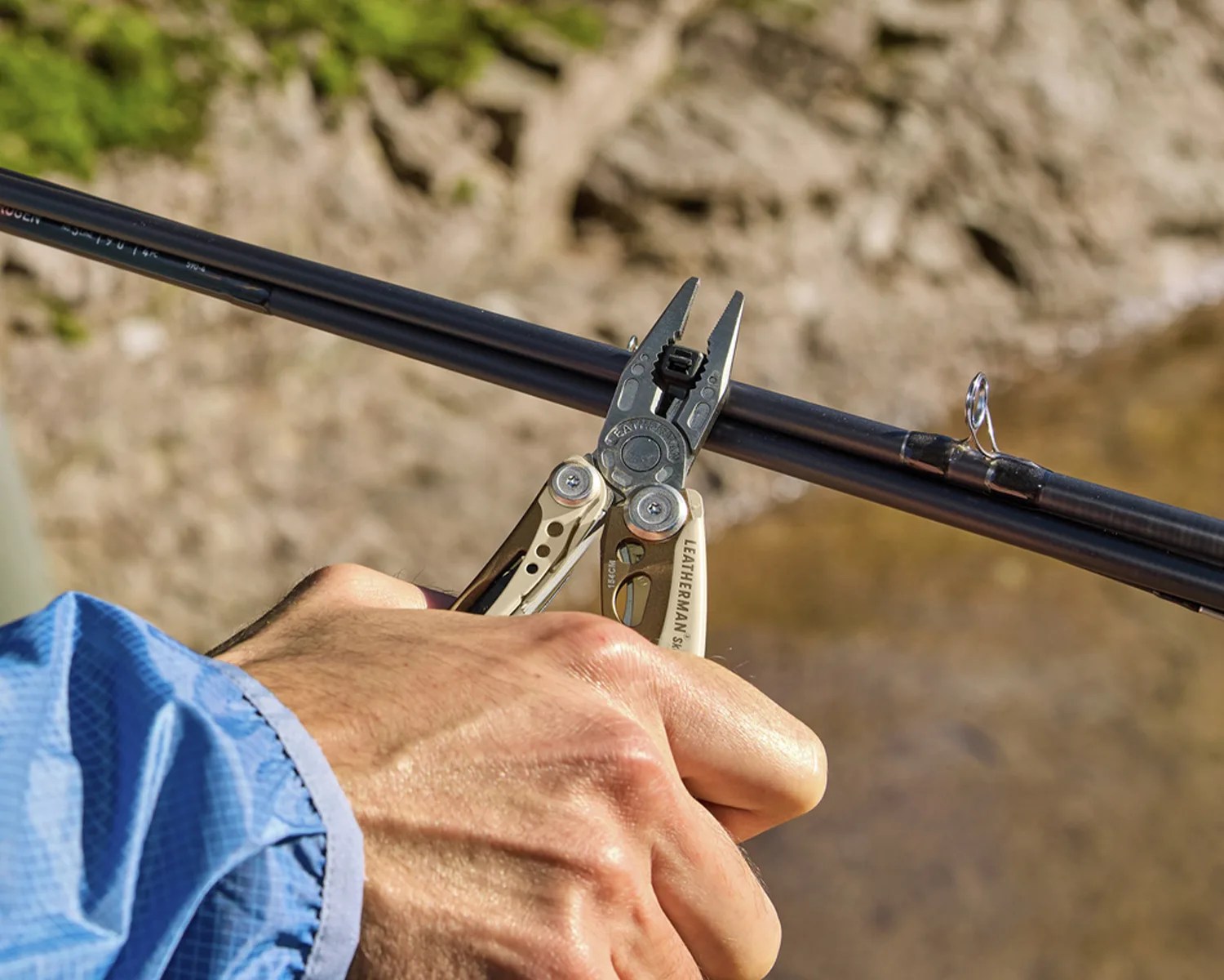 Hand holding a silver Leatherman multitool gripping a black fishing rod near a water body.