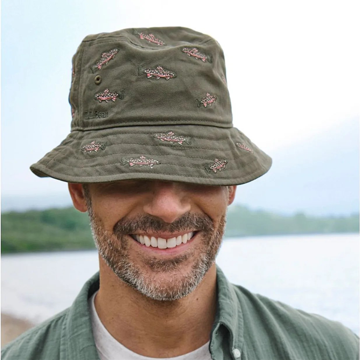 Olive-green bucket hat with embroidered pink and white fish worn by a smiling person outdoors.
